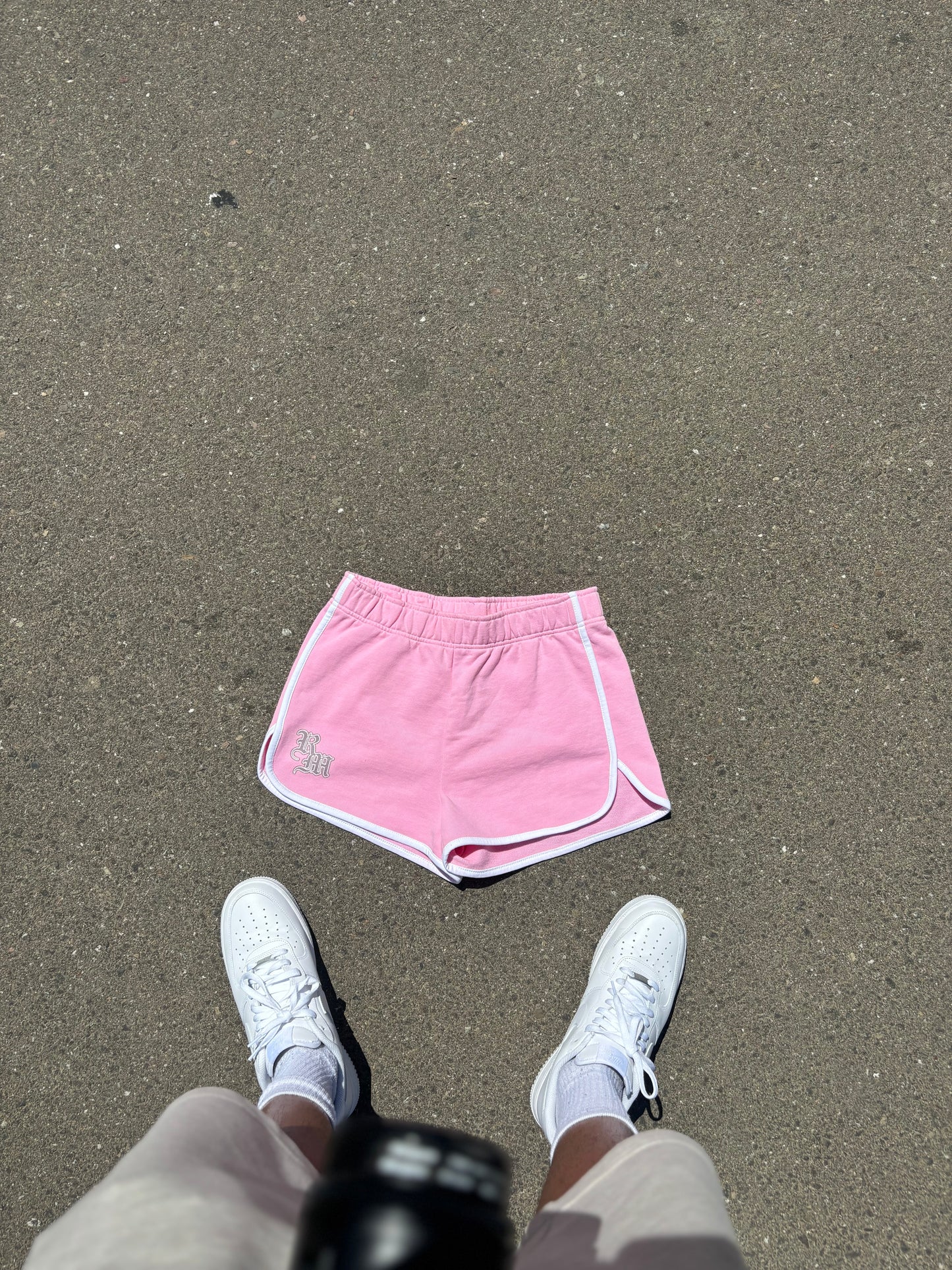Pink Renegade Mafia women’s shorts with white trim and RM logo on front, displayed on pavement with white sneakers.